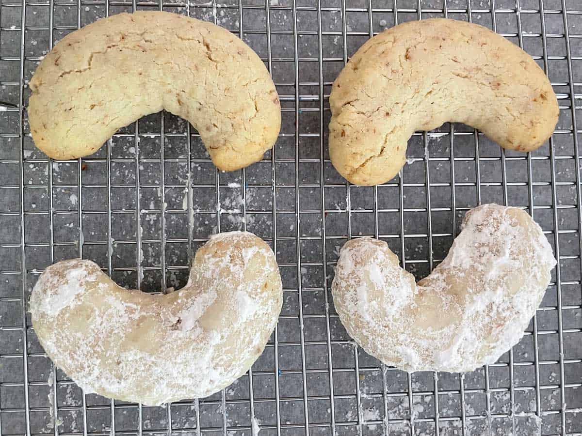 Four Almond Crescent cookies on a wire cooling rack. Two are finished and have been covered in powdered sugar, and two have not.