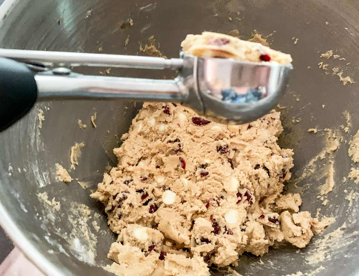 Cookie dough with cranberries and white chocolate chips being scooped out of a mixer bowl.