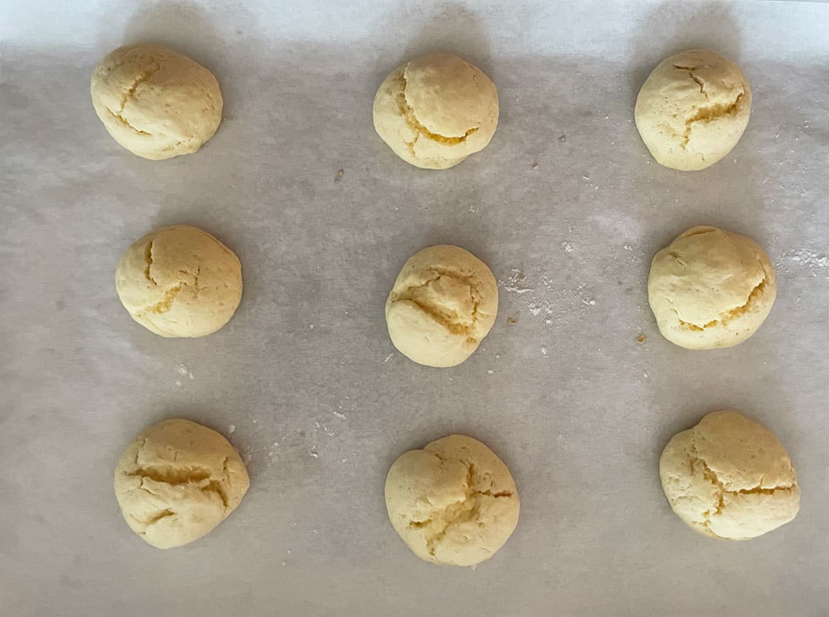 Nine balls of Italian Anise cookie dough on a parchment-lined cookie sheet pan after they are baked.