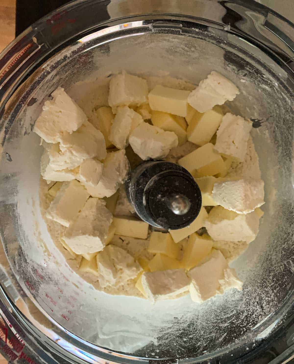 Flour mixture, slices of butter, and cream cheese pieces in a food processer bowl.