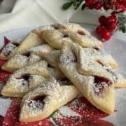 A stack of red raspberry kolache cookies on a Christmas plate with a sprig of red berries.