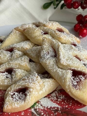 A stack of red raspberry kolache cookies on a Christmas plate with a sprig of red berries.