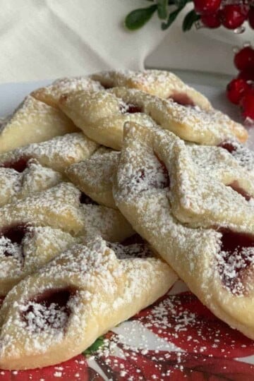 A stack of red raspberry kolache cookies on a Christmas plate with a sprig of red berries.