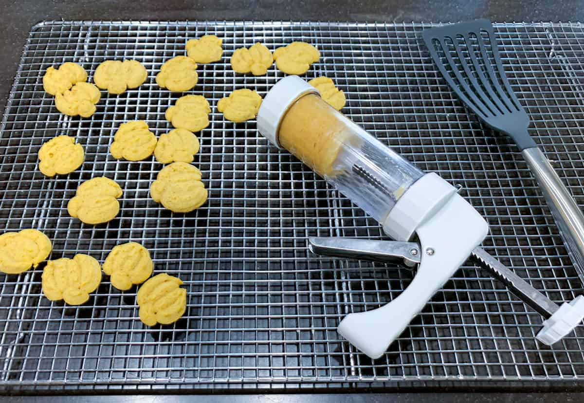 Cooling rack with a spritz gun, finished pumpkin spritz cookies, and a scooper to take cookies off a sheet pan.