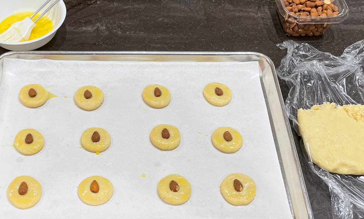 Cookie dough balls flattened and bushed with an egg wash before placing a whole almond on top.