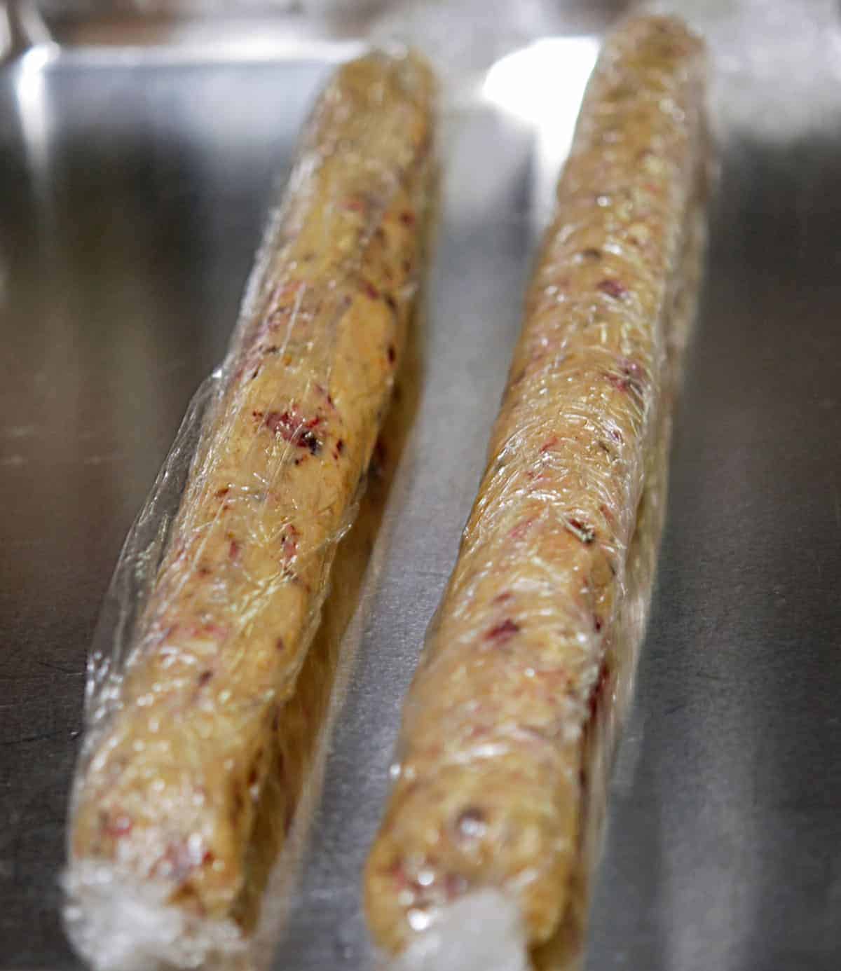 Two cookie dough logs wrapped in plastic wrap on a sheet pan and ready to go into the refrigerator.