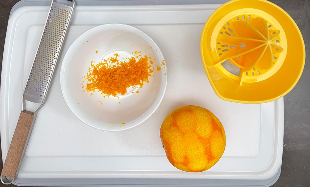 On a cutting board is an orange that has been zested, a zester, and a container for juicing the orange.