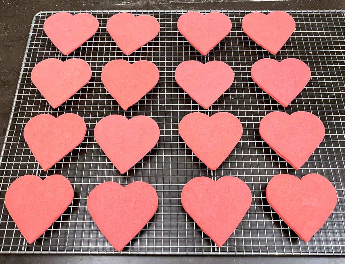 Sixteen finished pink heart sugar cookies cooling on a wire rack and getting ready to be decorated.