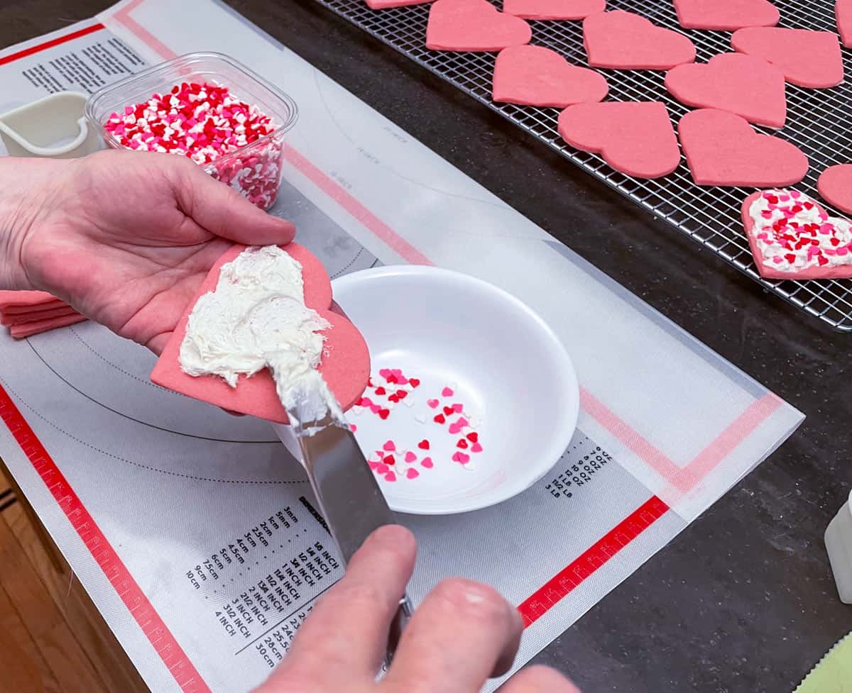 Using a knife to put buttercream icing onto a heart sugar cookie.