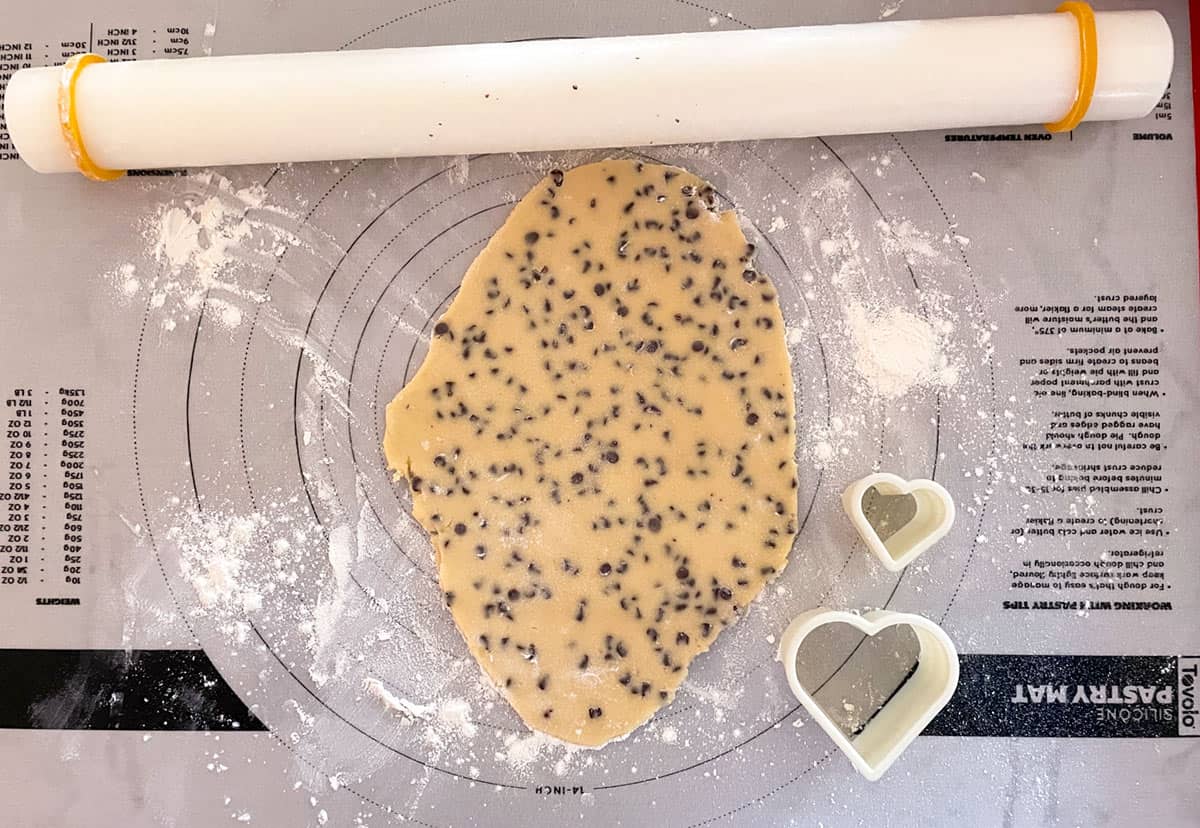 Cookie dough with chocolate chip rolled out on pastry sheet and two sizes of heart cookie cutters.