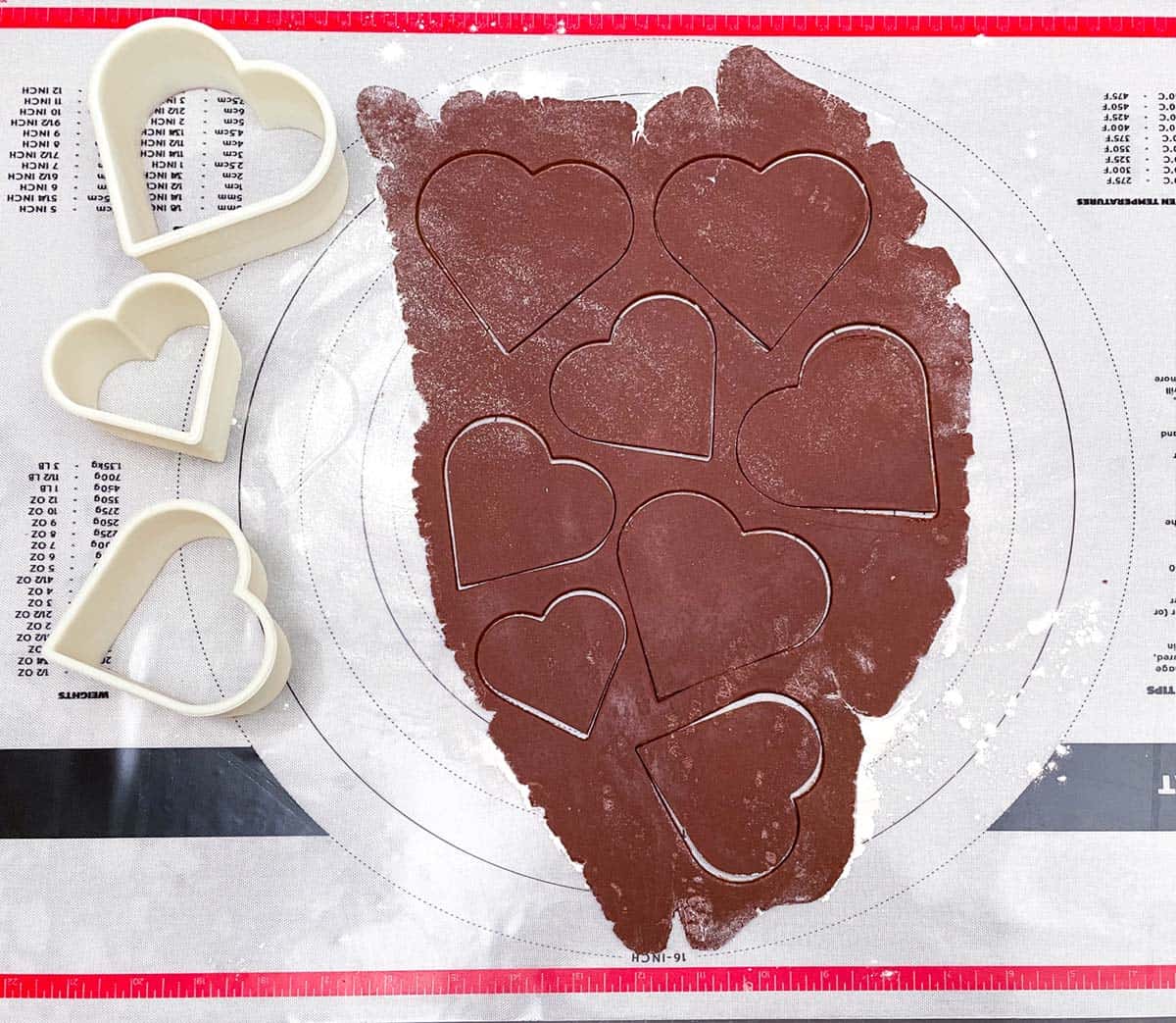 Three heart shaped cookie cutters with cut outs of rolled out chocolate cookie dough.