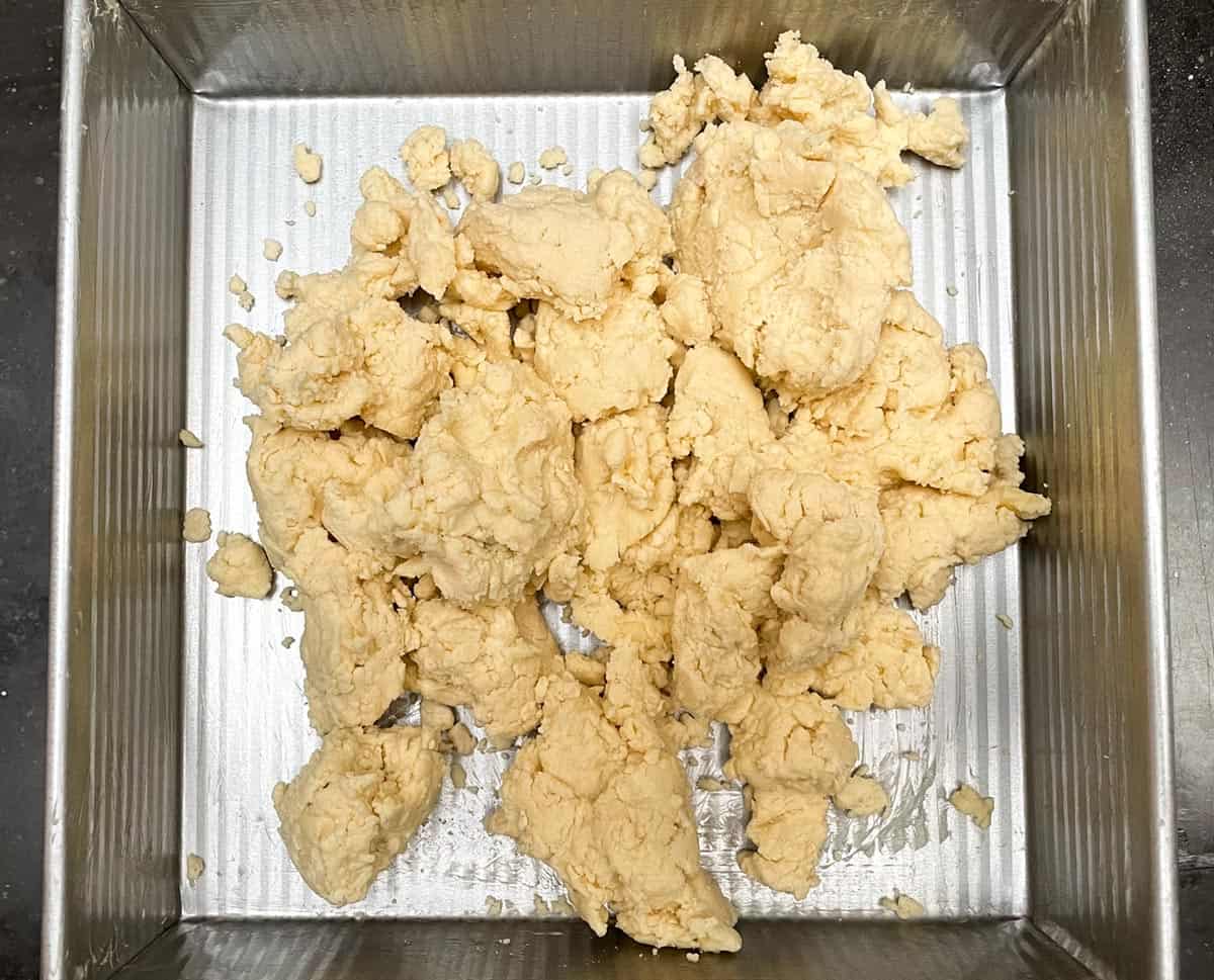 Clumps of shortbread dough that has been moved to a square baking pan.