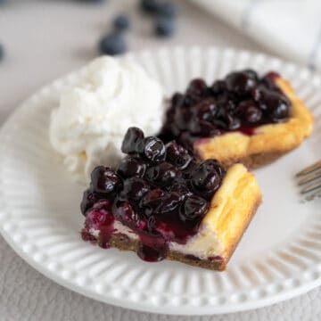 Blueberry Cheesecake bars on a white plate with blueberry sauce coming down the sides. A mound of whipped cream on the side.