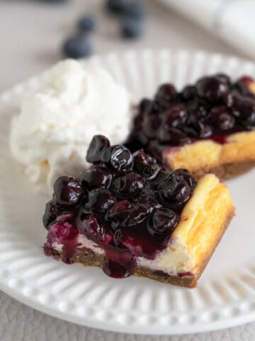 Blueberry Cheesecake bars on a white plate with blueberry sauce coming down the sides. A mound of whipped cream on the side.