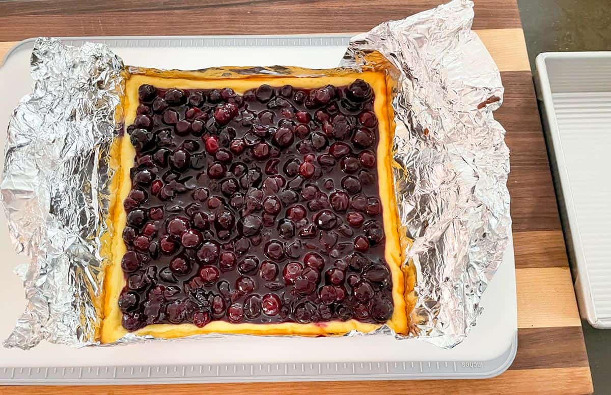 Square cheesecake with blueberry topping sitting on the tin foil liner from the baking pan.