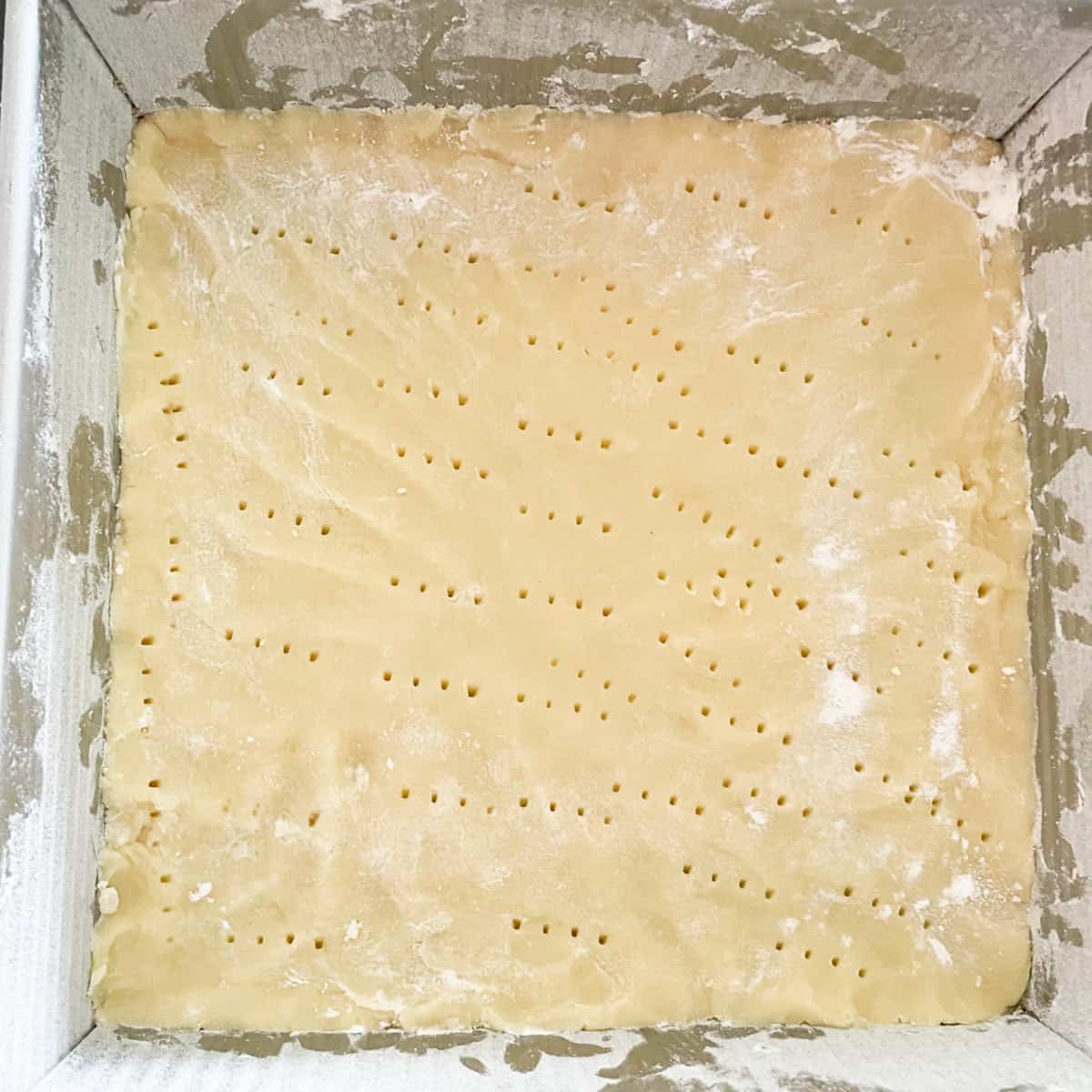 Shortbread crust pressed into square pan ready to be baked with fork marks.