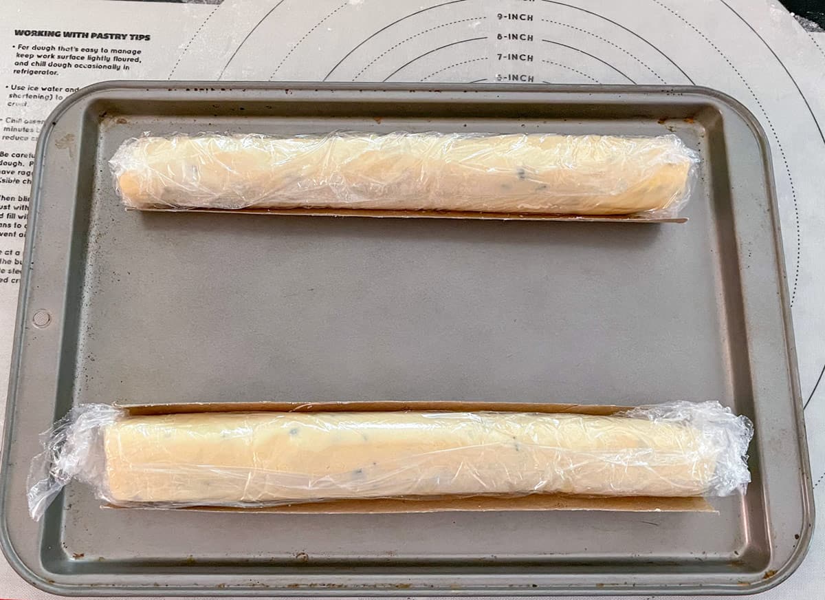 Two cookie dough logs wrapped in plastic and sitting in a mold to keep its shape.