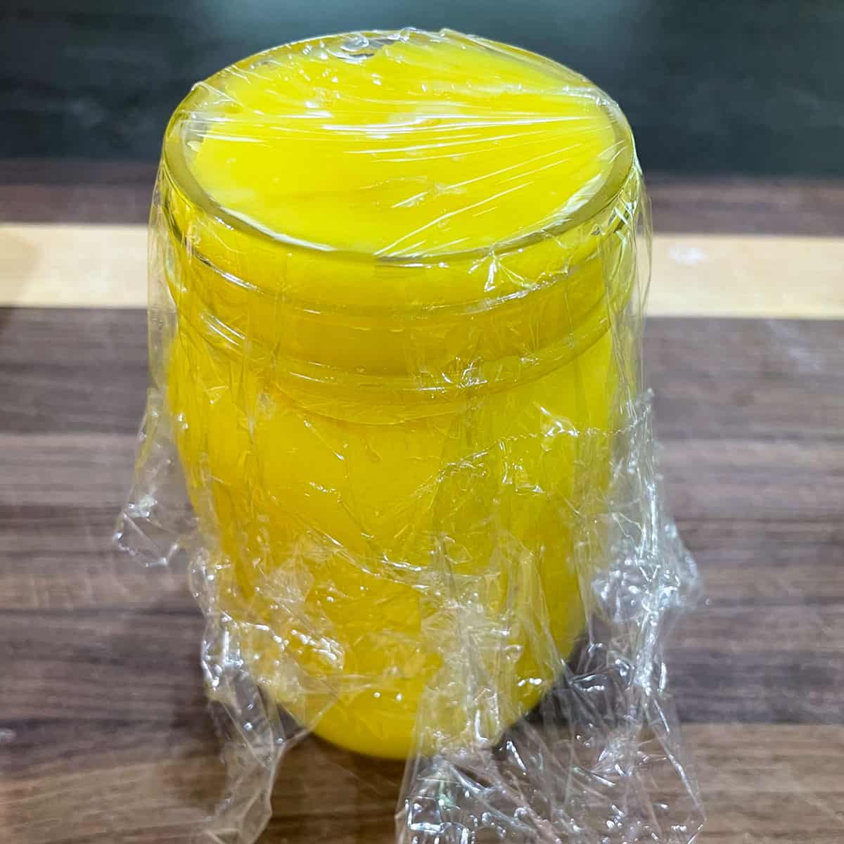 plastic wrap pressed against the curd that is in a glass jar.