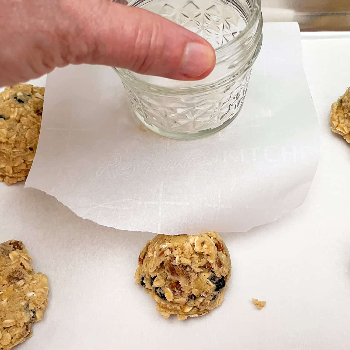 Trick for flattening a cookie mound using a square of parchment paper and a glass.