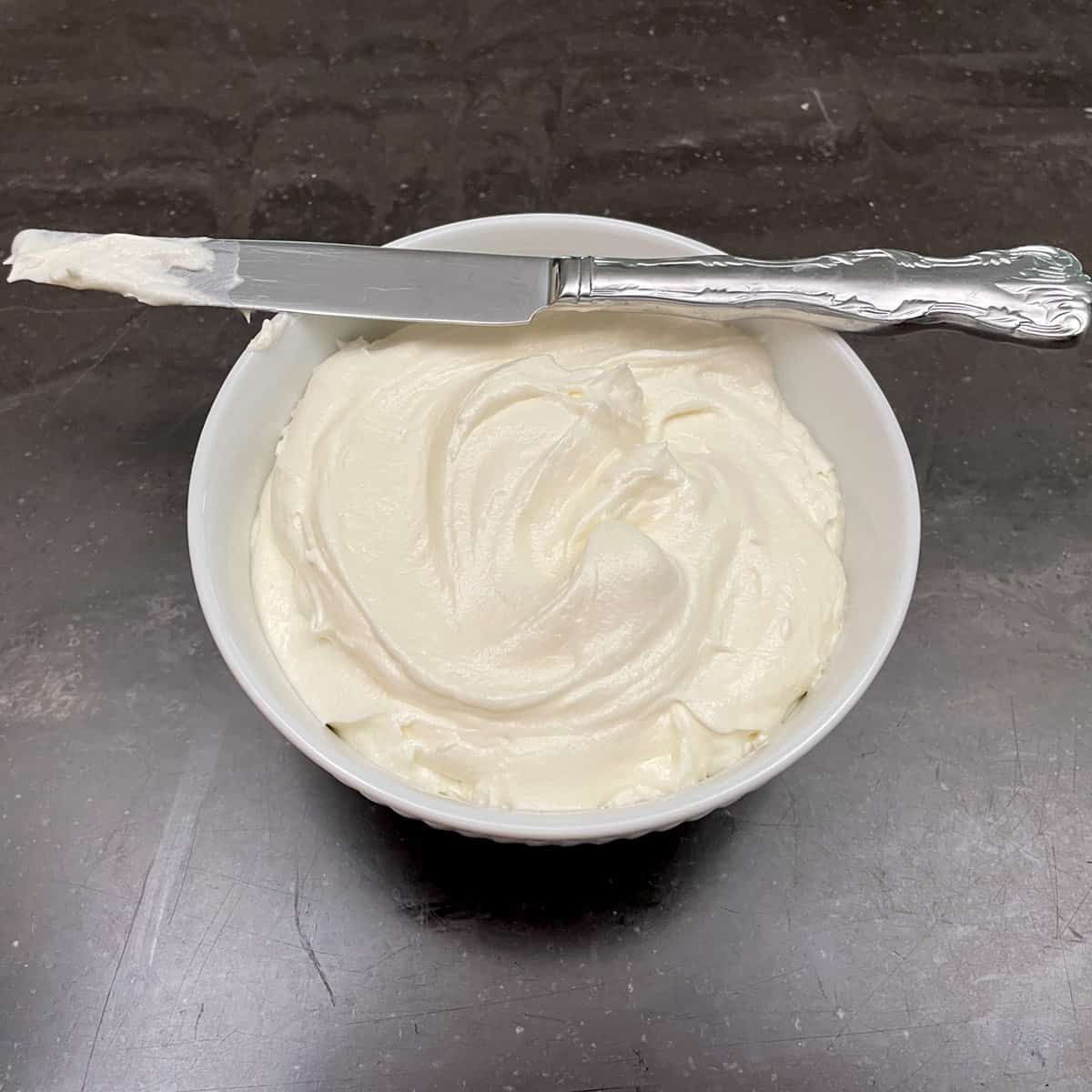maple cream cheese frosting in a white bowl with a knife laying across the top of the bowl.