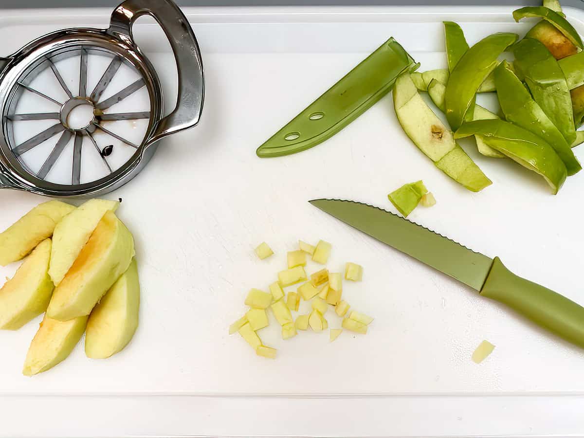 Cutting board with an apple corer, sharp knife, apple slices and apple peels.