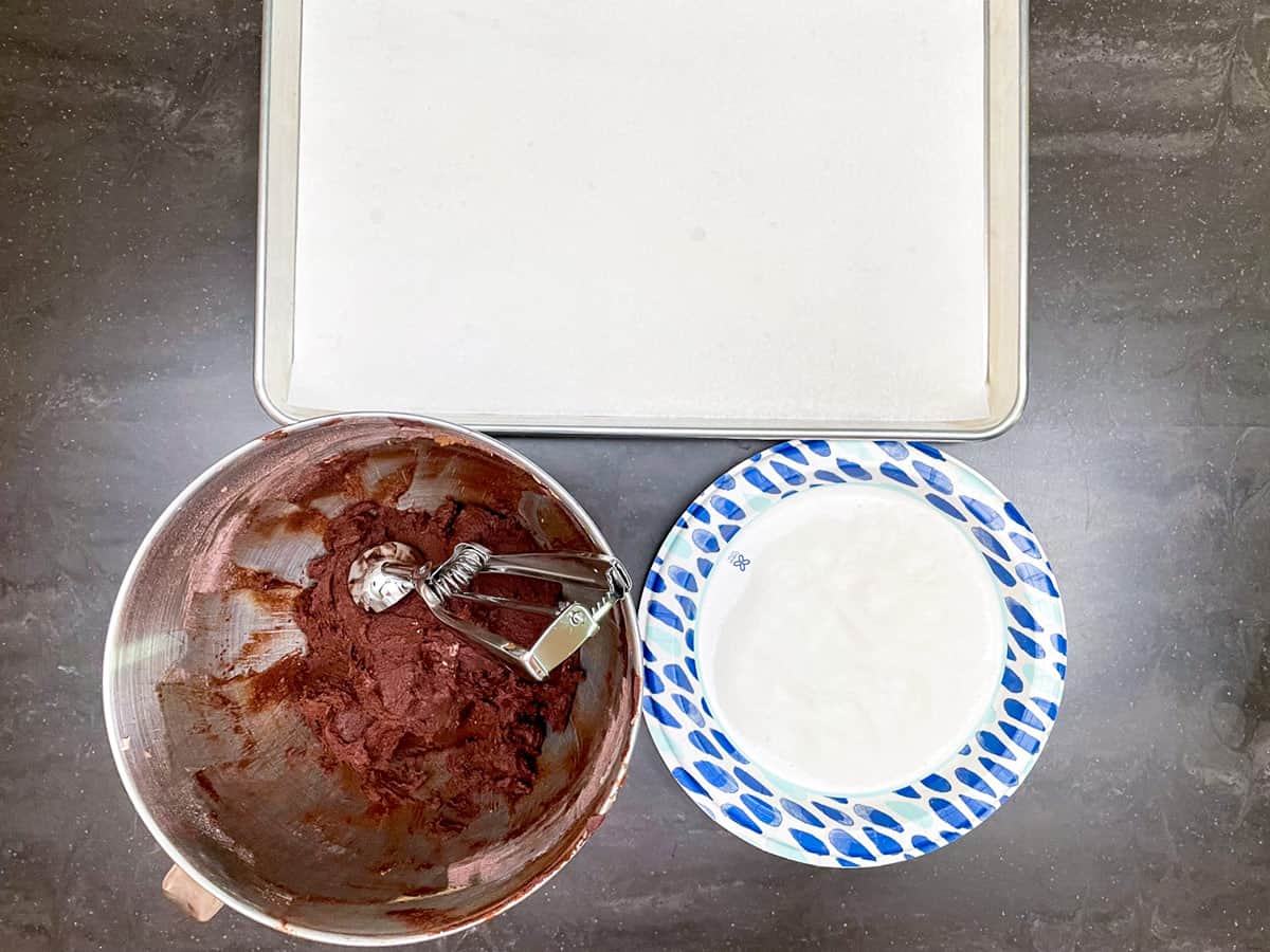 A bowl with cookies dough on left, a plate with sugar for rolling on the right and a parchment-lined sheet pan in the back.