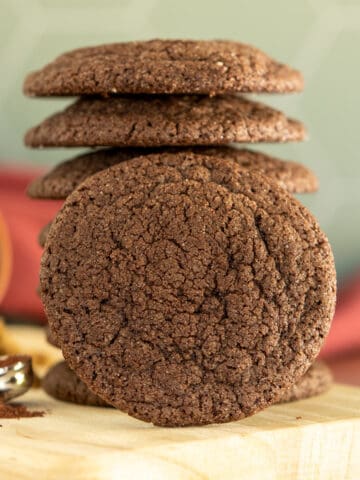 A stack of chocolate sugar cookies on a light-colored board with a raspberry cloth behind the stack.