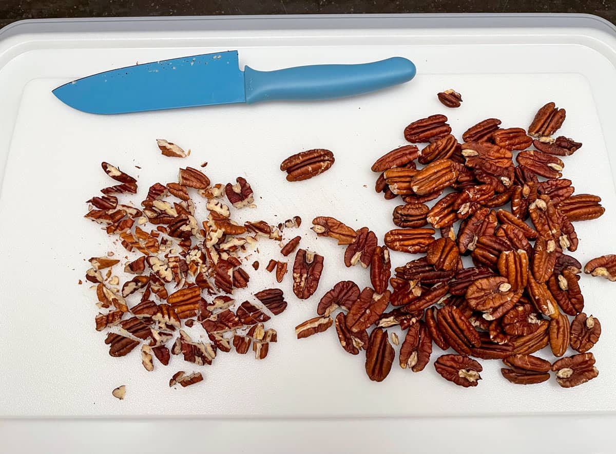 Rough chopping roasted pecans on a cutting board with a blue sharp knife.