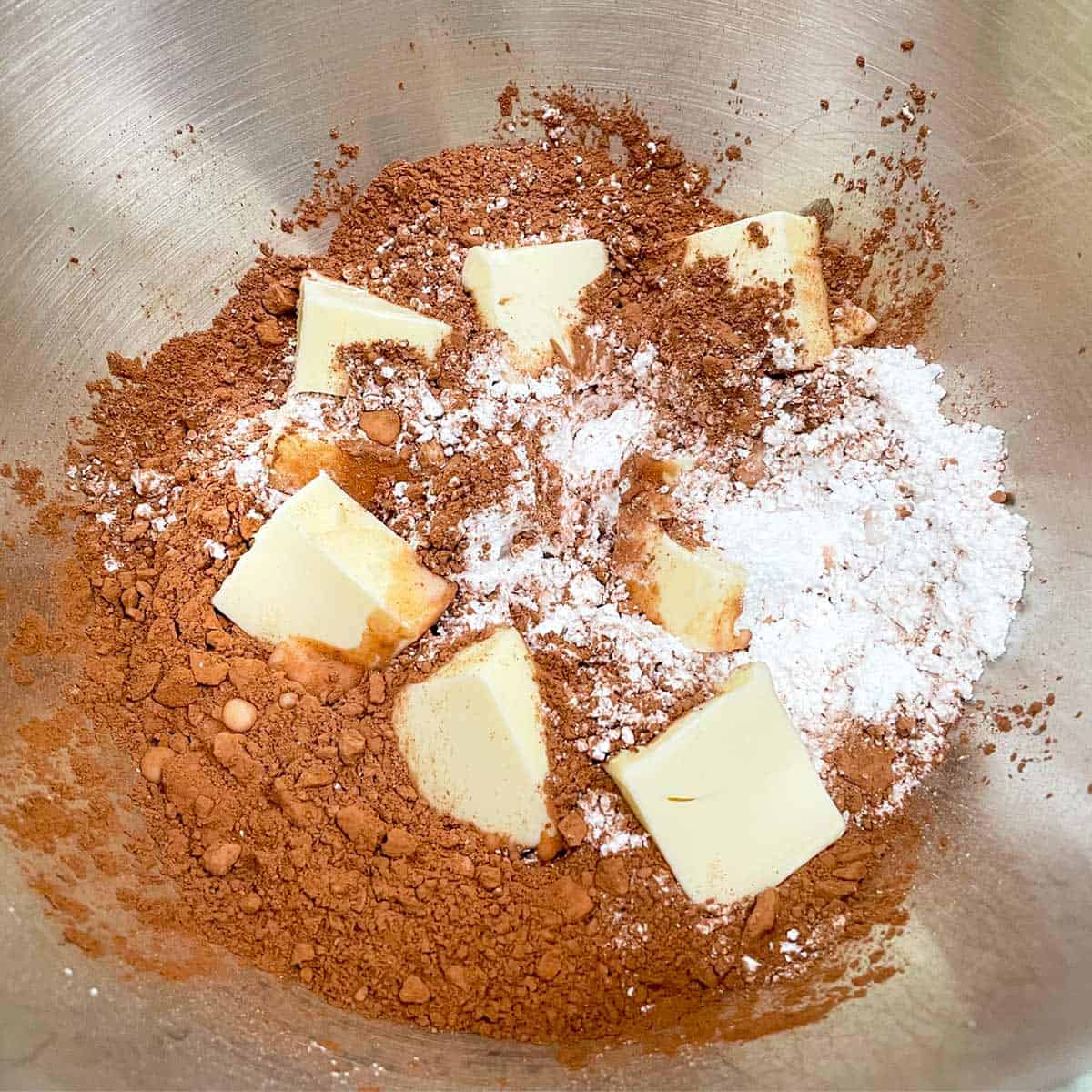 All ingredients to make chocolate icing in the mixer bowl including butter, cocoa, and powdered sugar.