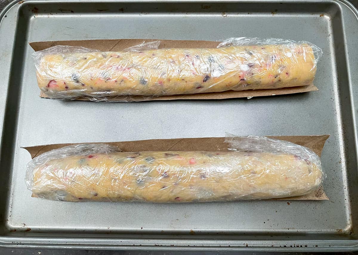 Cookie dough logs wrapped in plastic wrap sitting in cardboard cradles on a sheet pan ready for the chill.