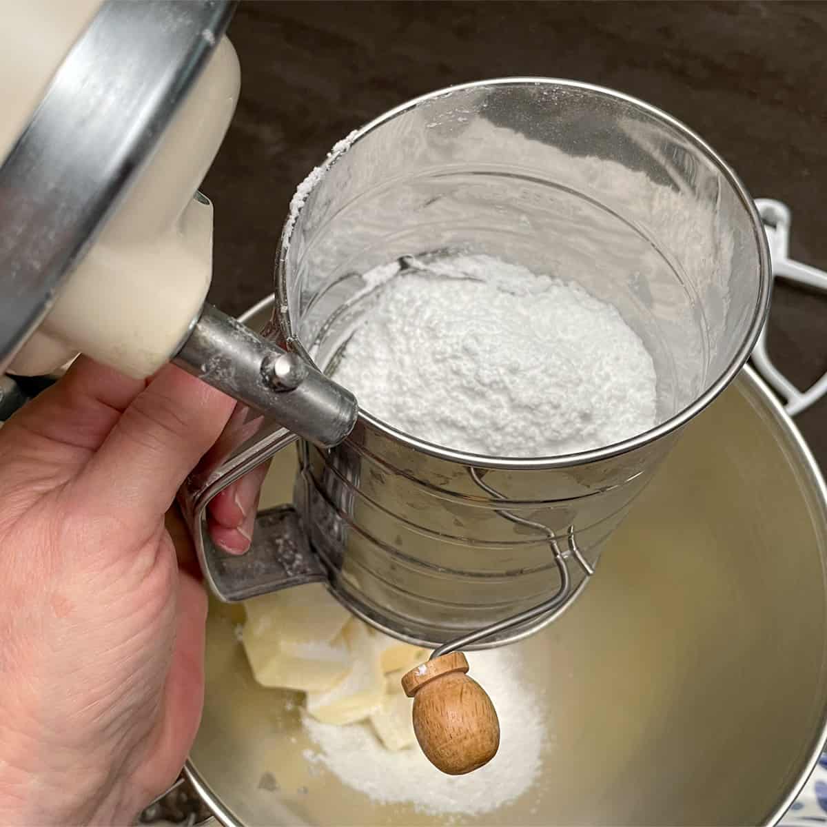 Sifting powdered sugar onto butter cubes in a mixer bowl.