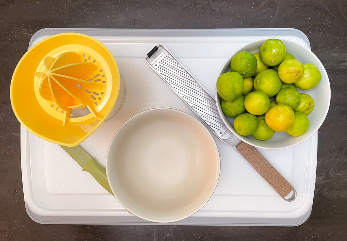 Cutting board with a bowl of key limes, zester, empty bowl, and a juicer.