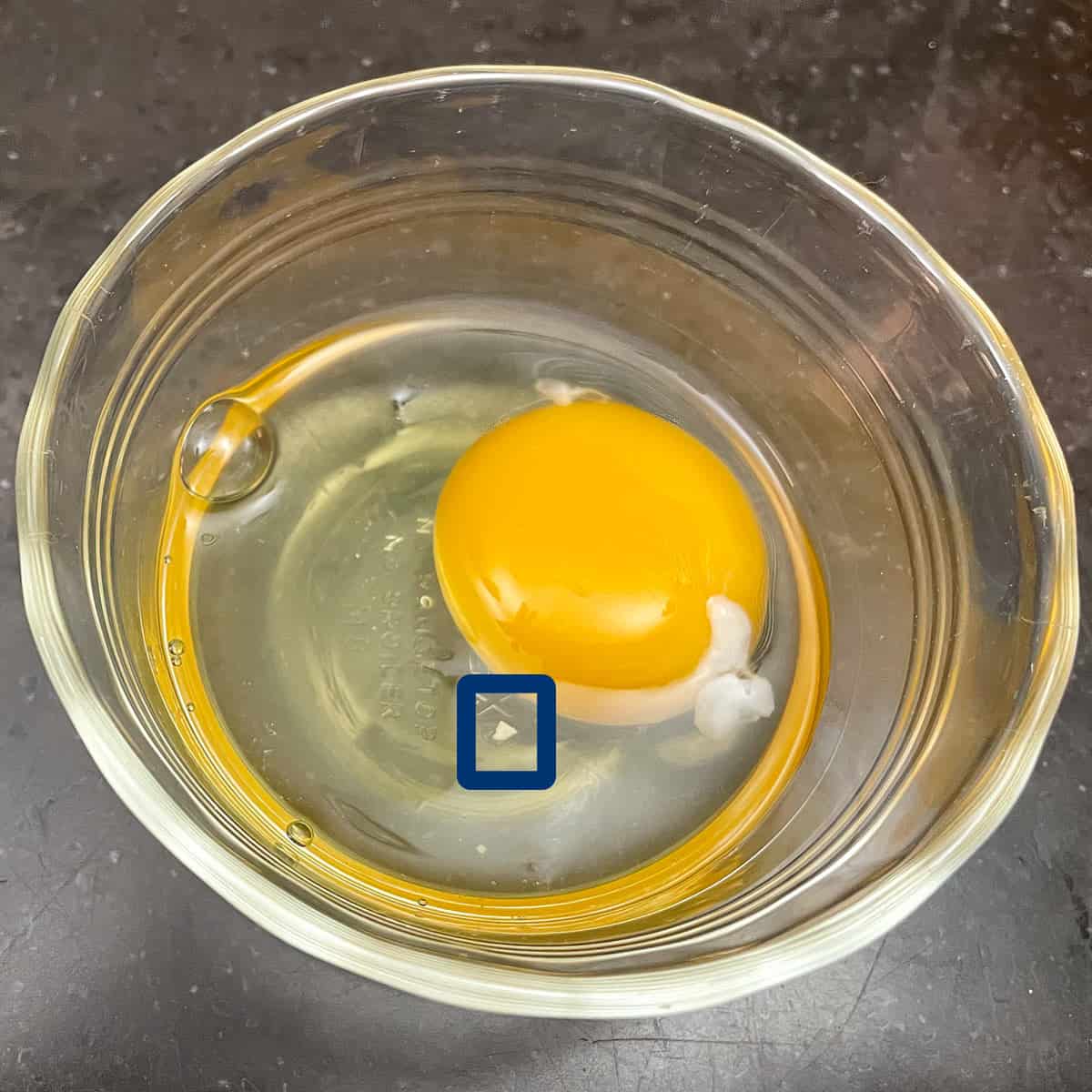 Raw egg in a small bowl showing a piece of eggshell that broke off when cracking the egg.