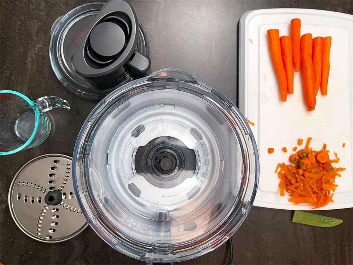 A food processor, measuring cup, a cutting board with whole trimmed carrots.