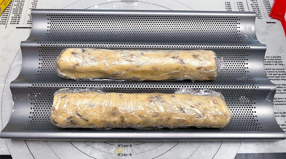 Two cookie logs wrapped in plastic wrap and laying in a pan that will keep them round.