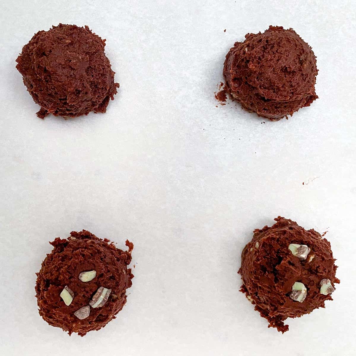 Four mounds of cookie dough where two of them have added mint chips to the tops before baking.