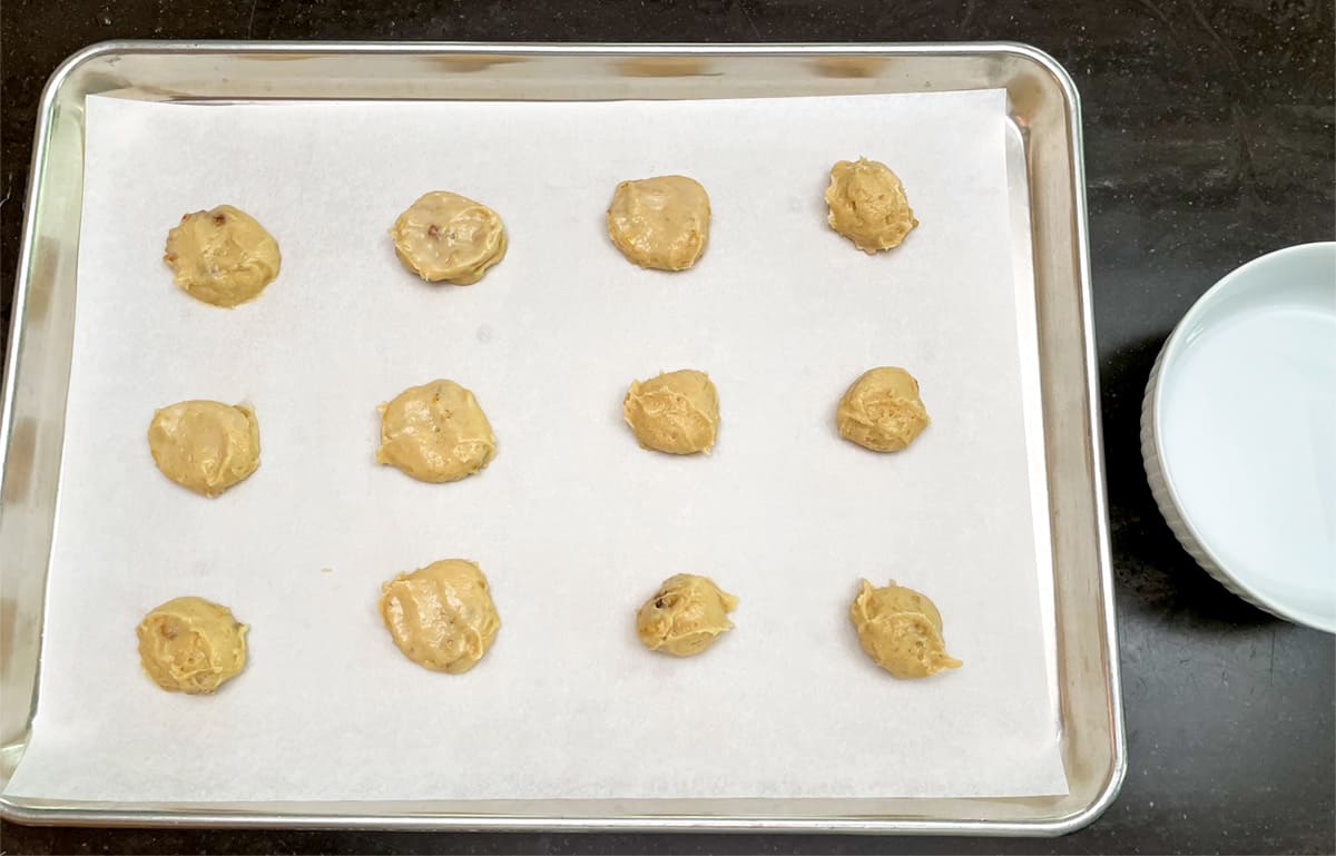 Twelve cookie mounds on a parchmentlined cookie sheet pan with a bowl of water next to it for wetting fingers.