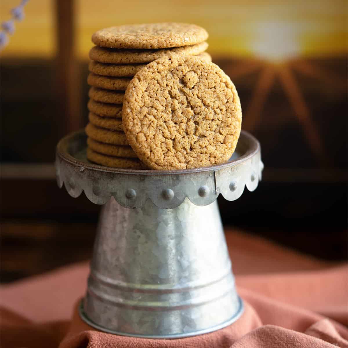 Stack of ginger cookies on a pewter stand with a window scene behind it.