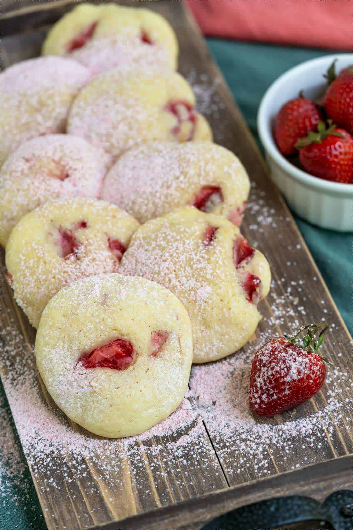 Display a group of strawberry shortcake cookies on a long board with fresh strawberries on the side.