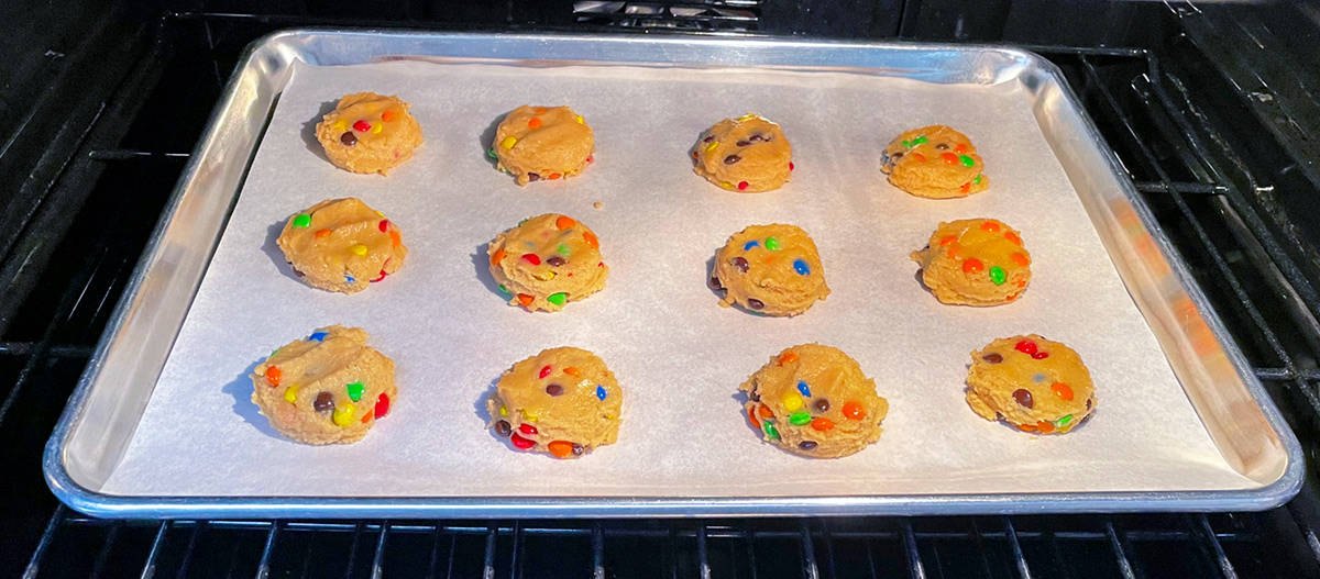 Twelve scooped Peanut Butter M&M cookie dough on a sheet pan in the oven that have been slightly flattened and shaped around the sides.