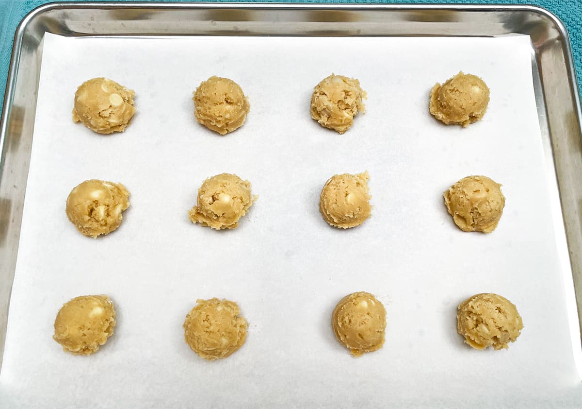Twelve white chocolate macadamia nut cookie dough scooped onto parchment paper that is on a sheet pan.