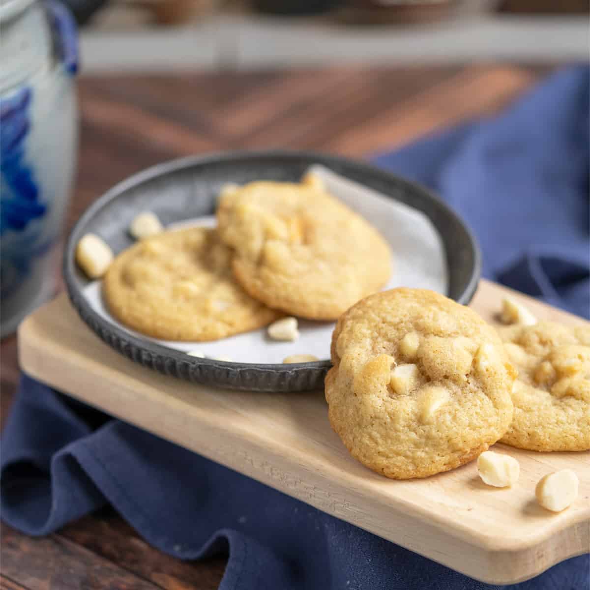 Four white chocolate macadamia nut cookies on a wooden board with a towel under it.