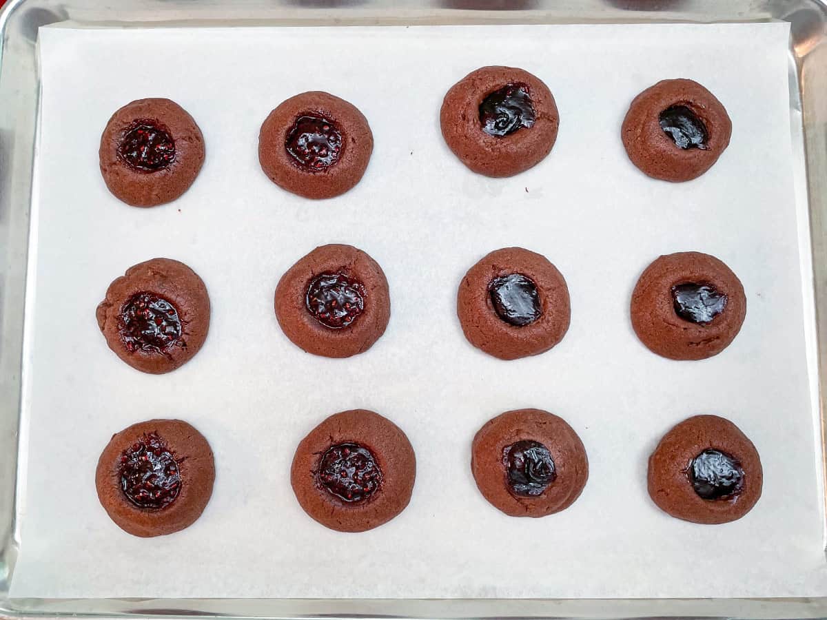 Twelve prebaked chocolate thumbprint cookies with raspberry jam sitting on a parchment lined sheet pan.