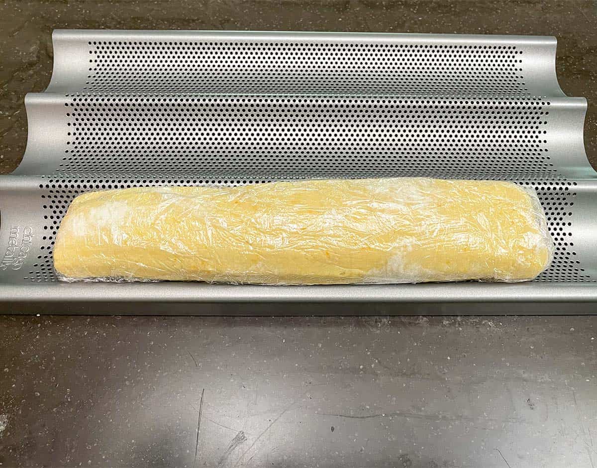 Cookie dough log wrapped in plastic wrap and resting in a baguette pan ready to be chilled.