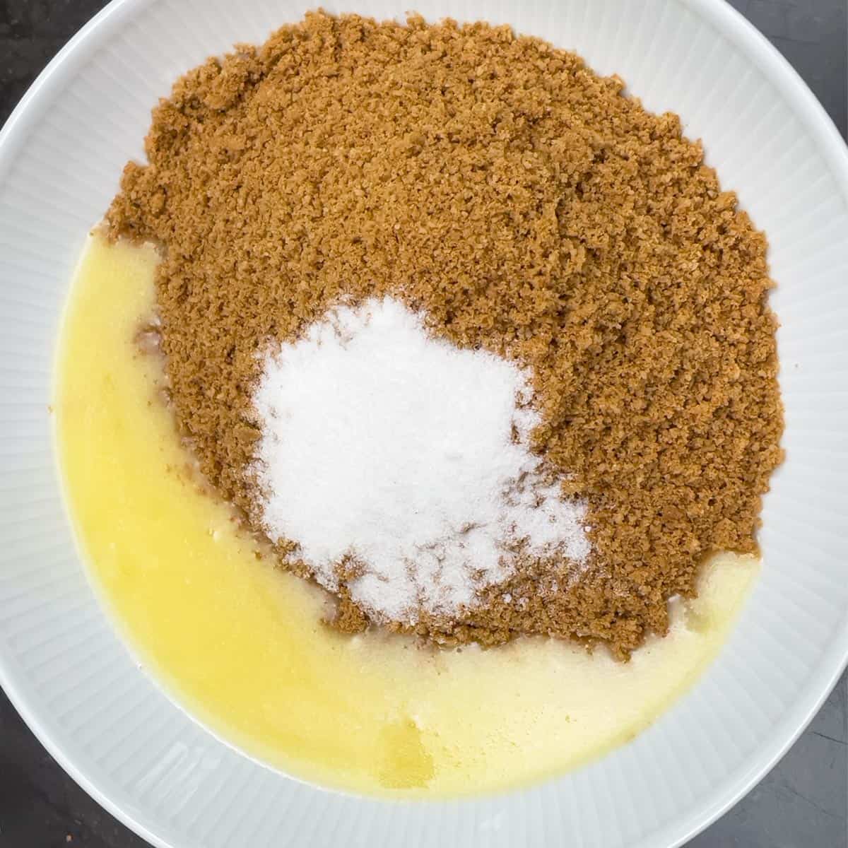 Graham cracker crumble in a dish along with melted butter and sugar to be mixed.