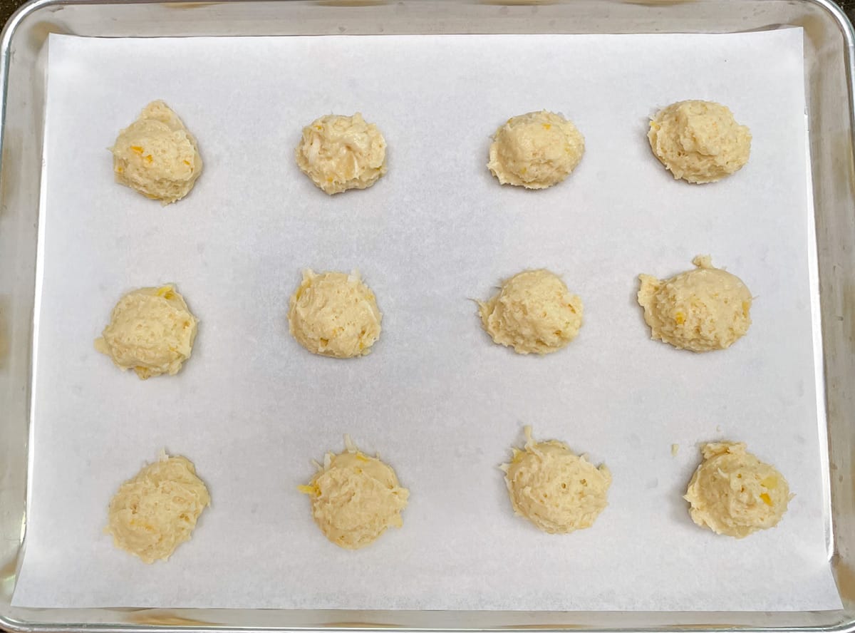 Twelve scoops of pineapple coconut cookie dough on a parchment lined cookie sheet pan.