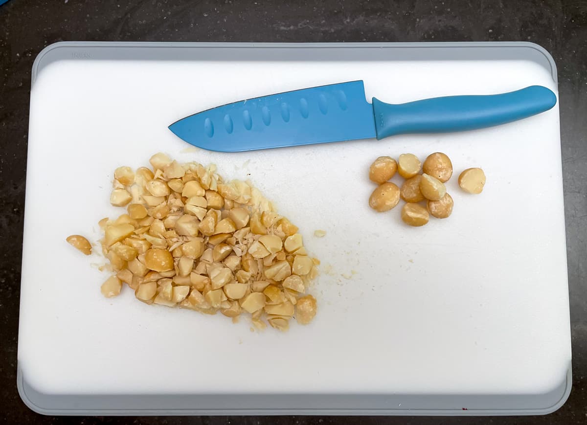 Chopping up roasted macadamia nuts on cutting board with sharp knife.