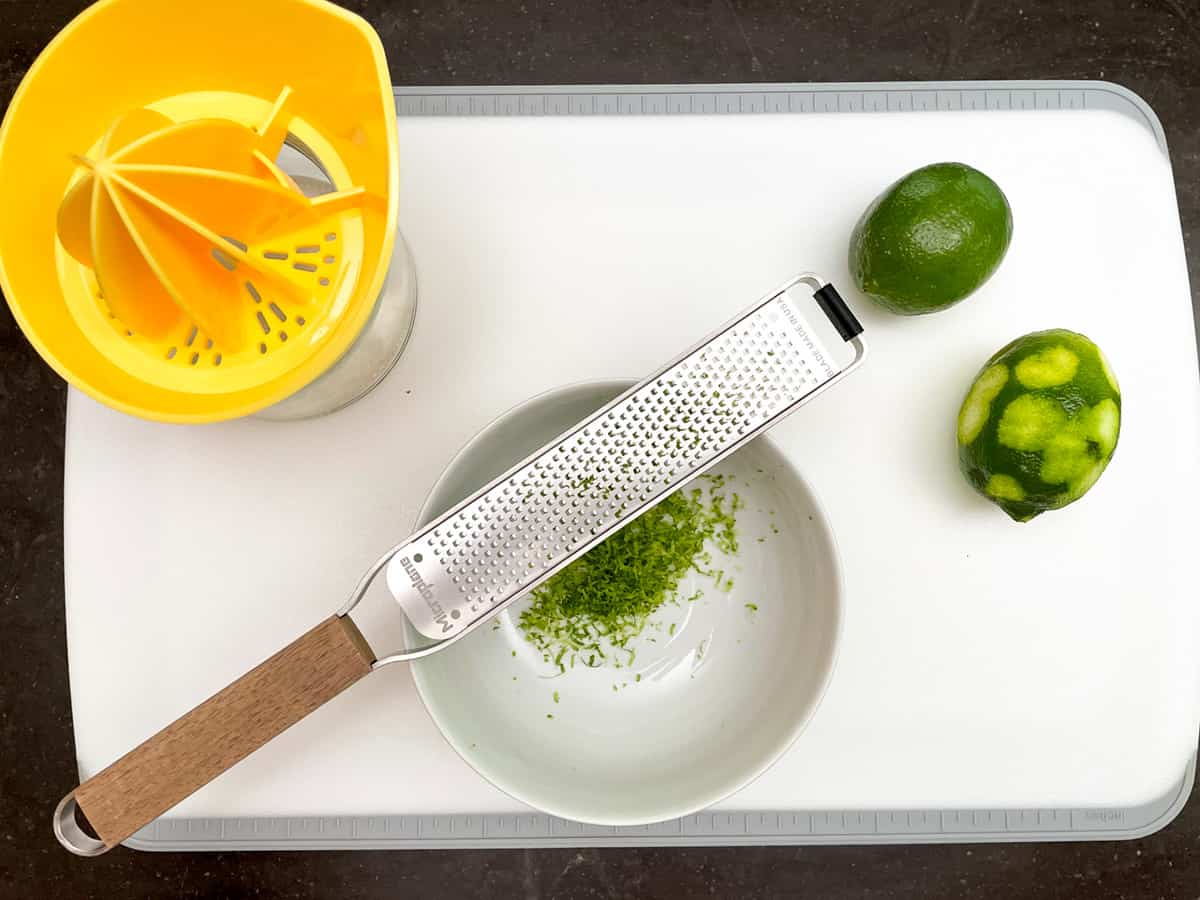 Zesting and juicing limes on a cutting board with a bowl, zester and juicer.