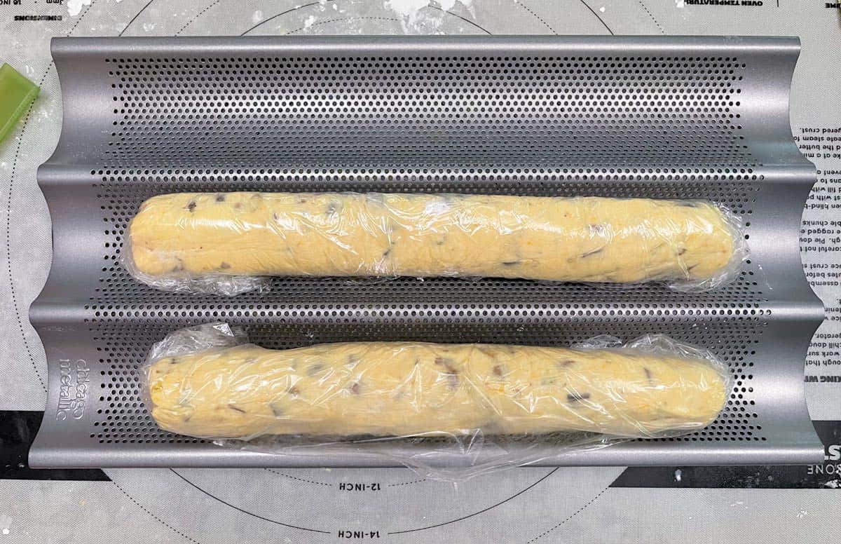 Two cookie logs that are wrapped in plastic and resting in the curves of a baguette pan.