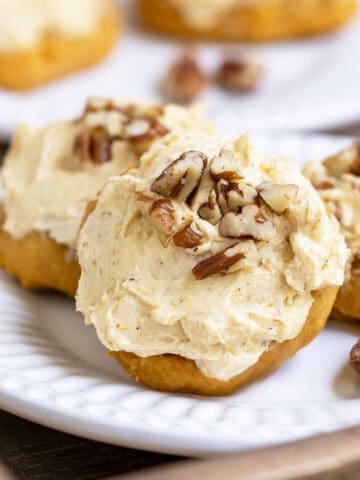 All about pumpkin cookies on a plate. Cookie with frosting and chopped pecans on top.